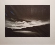 Black and white print of riverbend. Most light is at center of horizon which illuminates scattered clouds just above the river and shimmers light off the river to show us where it flows.